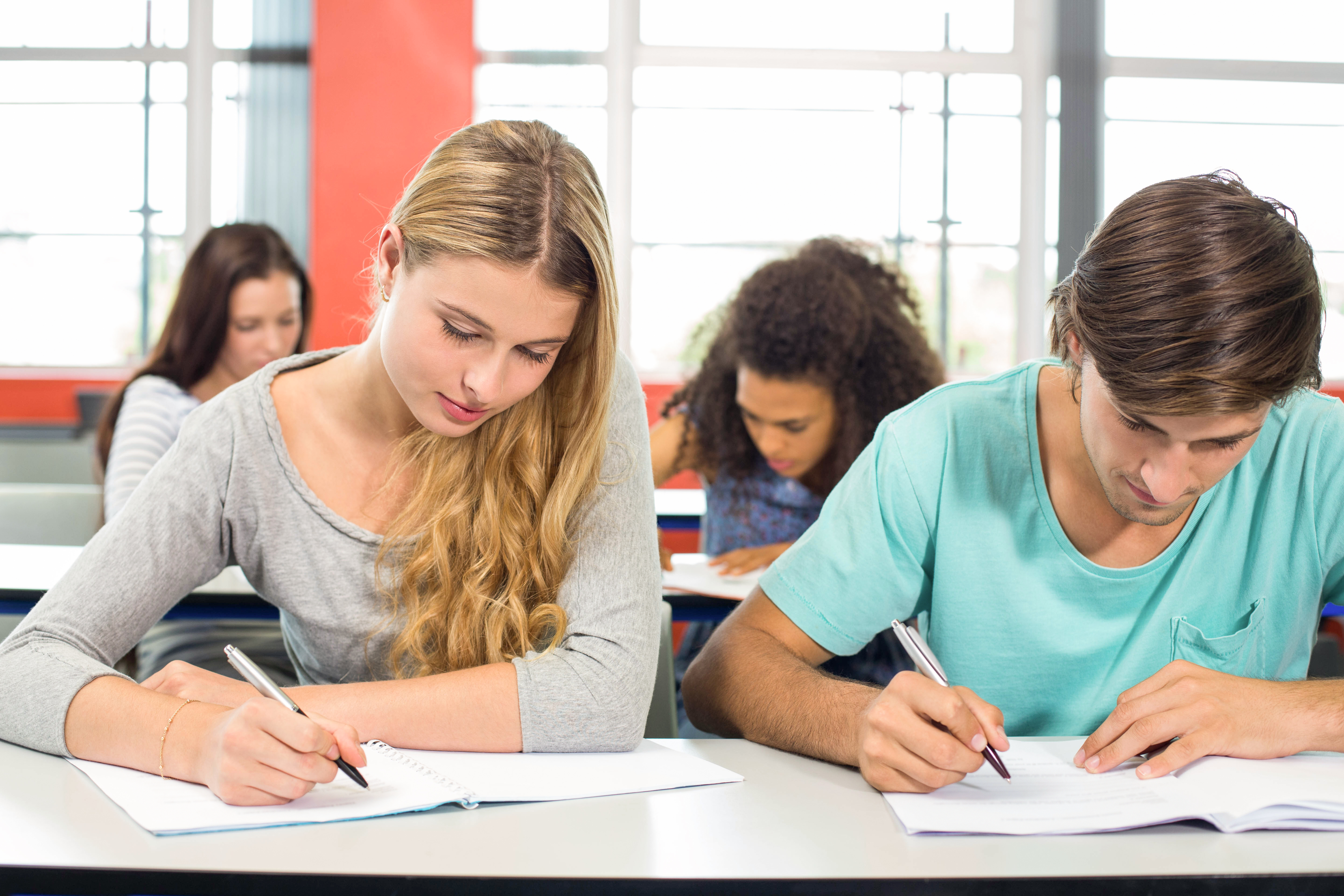 Two students sitting next to each other and writing, with two more students in the background.