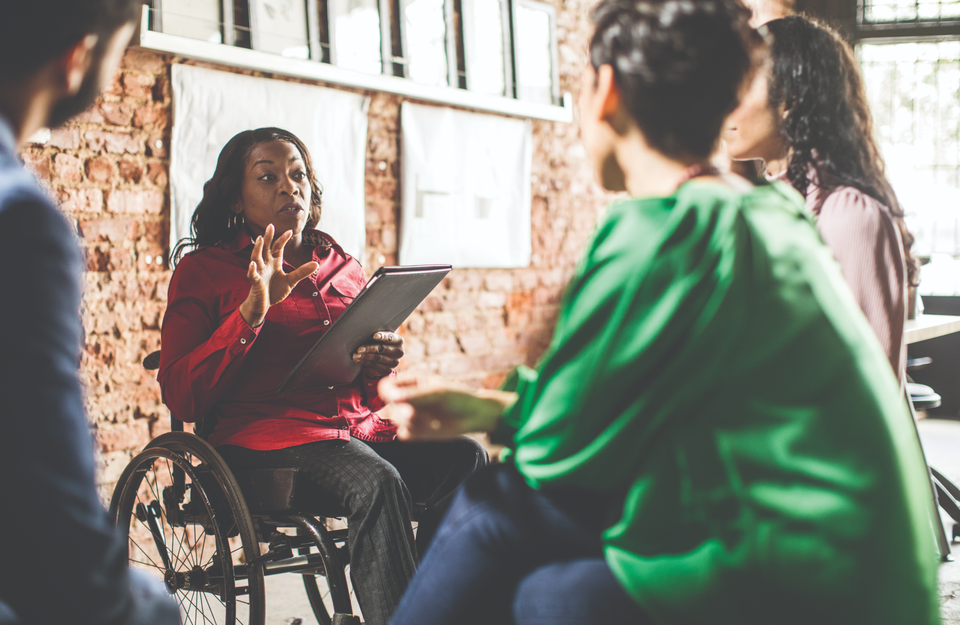 A woman leads a meeting in a brick-walled room while three attendees look on.